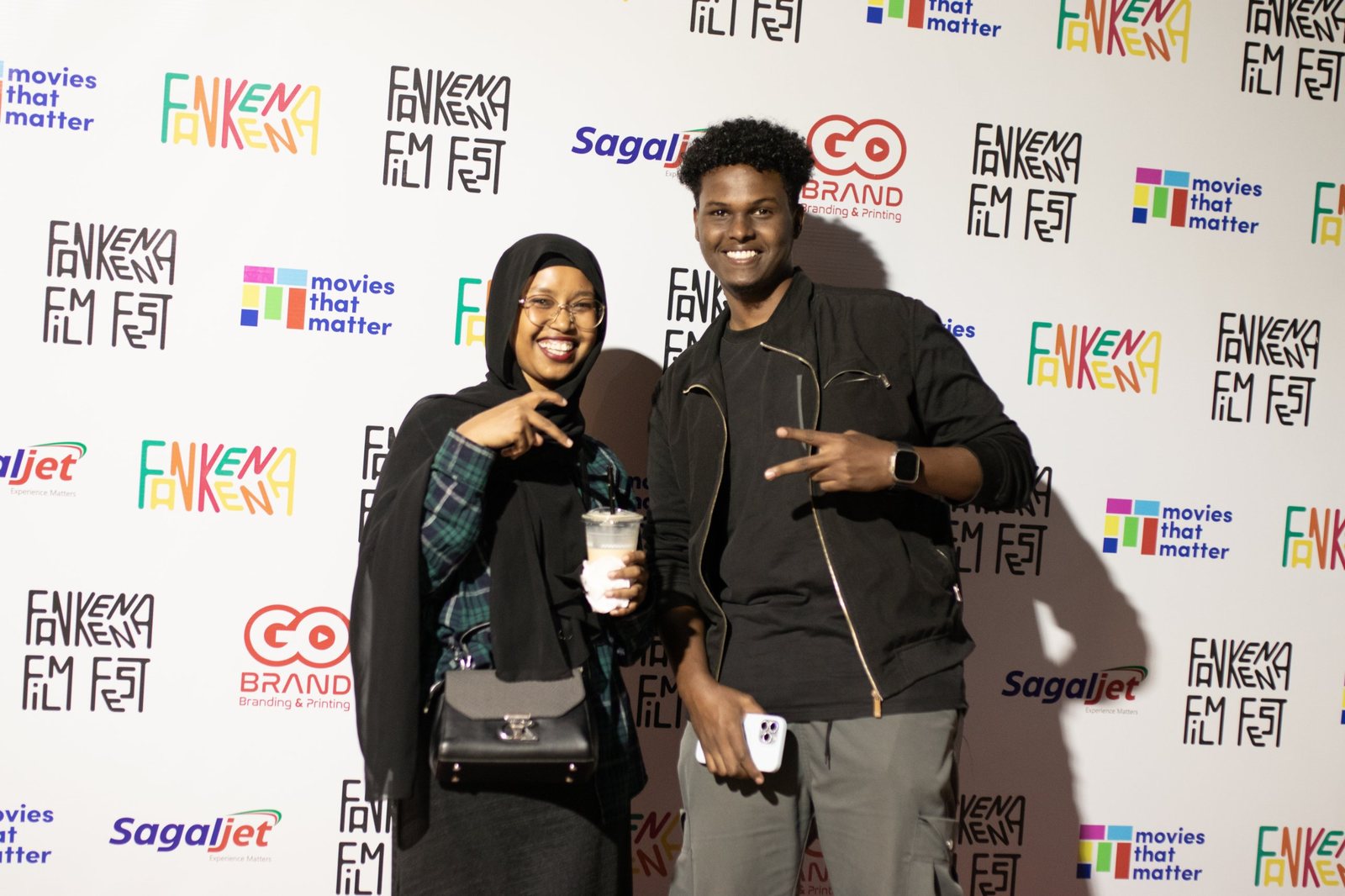 Two guests smile on the red carpet in front of the sponsor wall